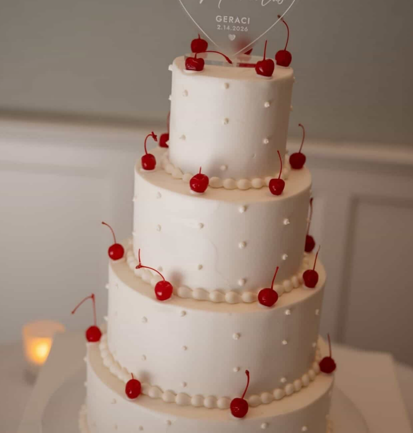 A four-tiered white wedding cake features small white dots around the layers and red cherries on top of each layer.