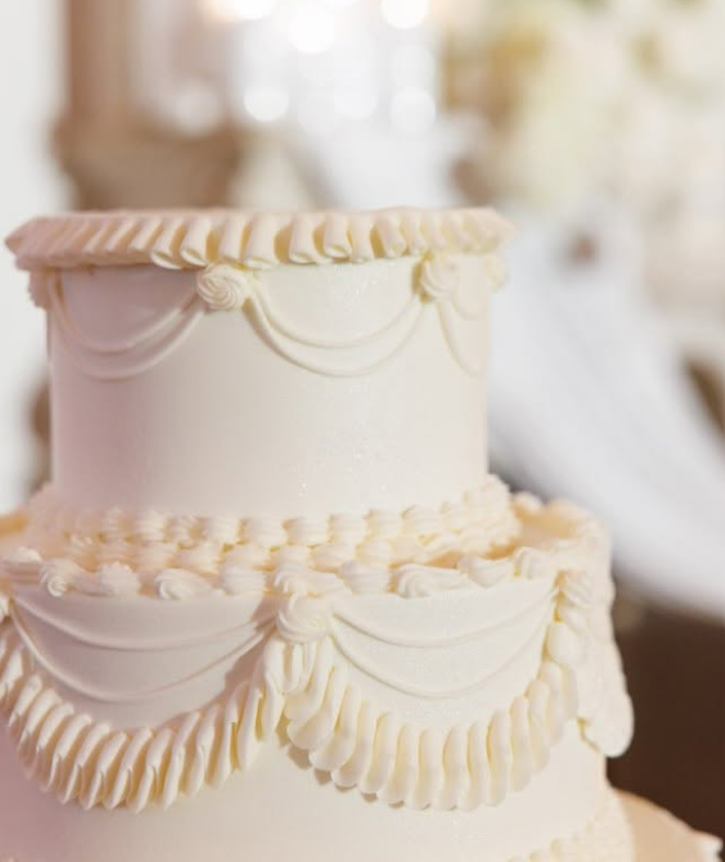 A close-up view shows two layers of a white cake that features custom piping around the edges. The background is blurry.
