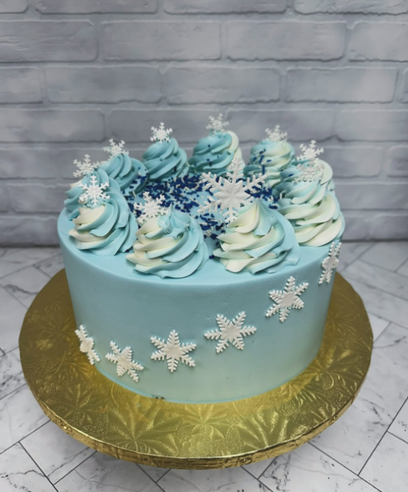 A round light-blue cake has white snowflakes around the bottom and white and blue icing swirls on top.