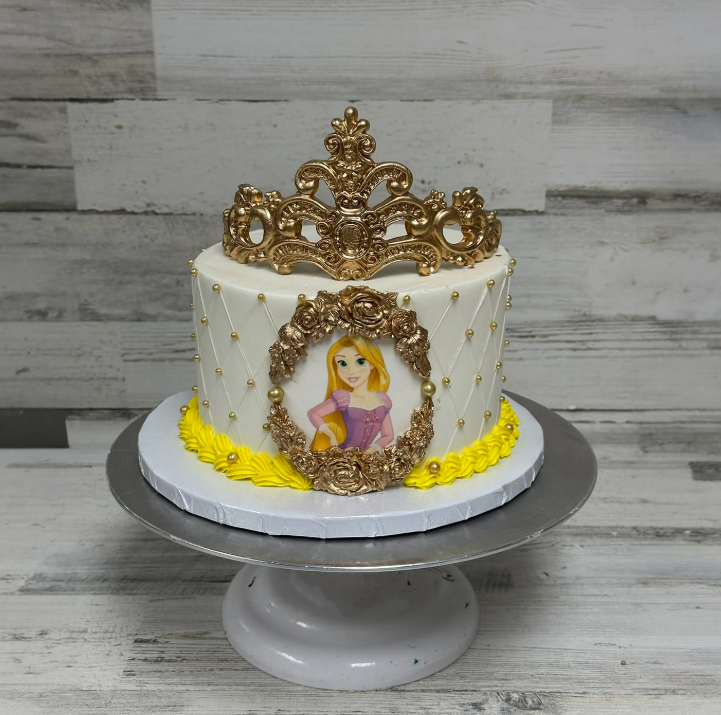 A round white cake has yellow icing swirled around the bottom, a photo of Rapunzel, and a gold crown on top.